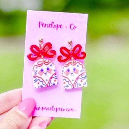 A hand holding a pink card with earrings on it, against a blurred green background.