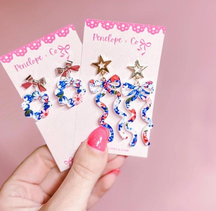 A hand holding two pairs of earrings with floral designs and bows.