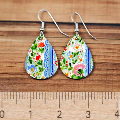 A pair of colorful earrings with a floral pattern, placed on a wooden surface next to a ruler for size comparison.