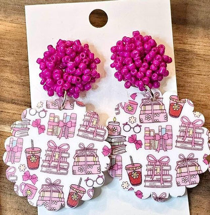a pair of earrings with a pink bead design and a scalloped paper pattern featuring various pink and white illustrations, such as gift boxes, flowers, and other decorative elements.