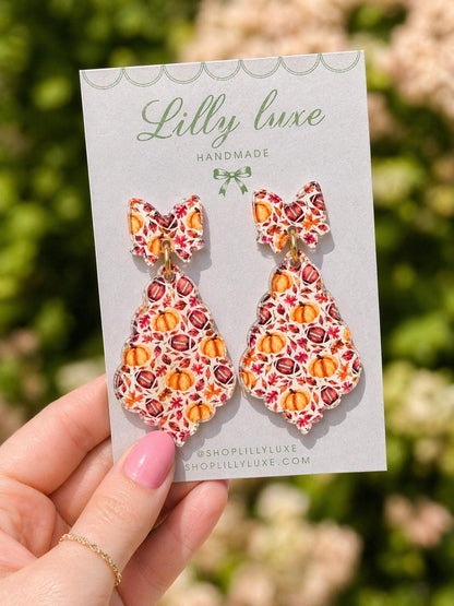 A hand holding a card with earrings featuring a pumpkin and leaf pattern, against a blurred background of greenery.