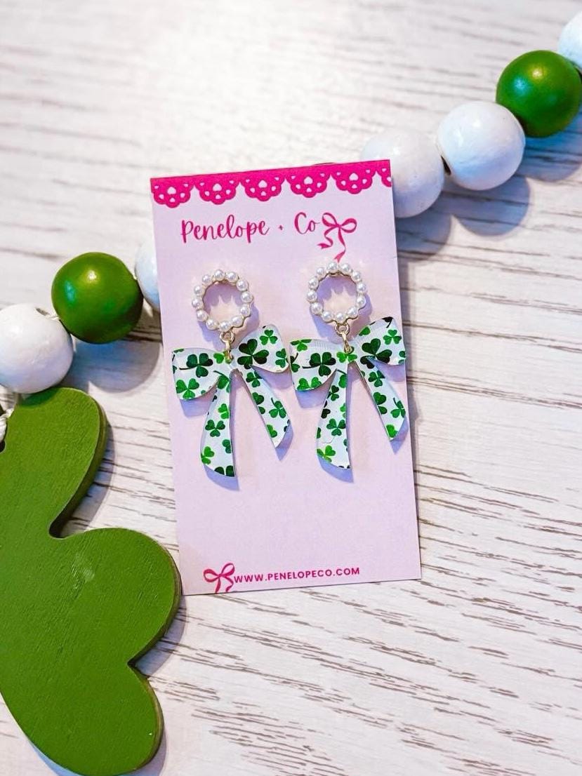 A pair of earrings with green bows and white beads, displayed on a pink card with a website address.