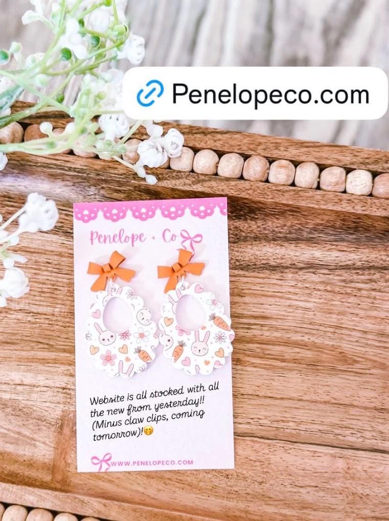 a pair of earrings with bows, placed on a wooden surface. The earrings are described as "Mini claw clips" and "coming tomorrow". The background includes a floral arrangement and a website address for Penelopeco.com.