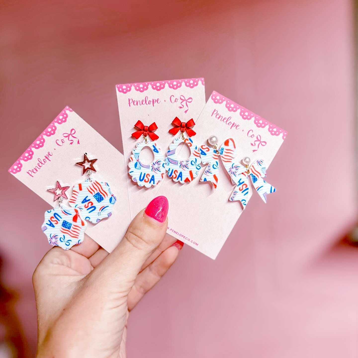 A hand holding three earring cards with patriotic designs, including stars, stripes, and the word "USA".