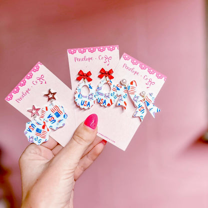 A hand holding three earring cards with patriotic designs, including stars, stripes, and the word "USA".